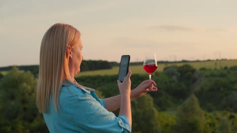 A-woman-blogger-photographs-a-glass-of-wine-against-the-backdrop-of-a-picturesque-valley-where-the-sun-sets