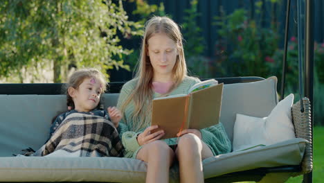 A-teenage-girl-reads-a-book-to-her-younger-sister.-Resting-together-on-a-garden-swing-in-the-backyard-of-the-house