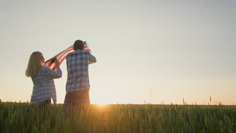 Celebrating-Independence-Day---a-man-is-waving-the-US-flag,-a-woman-nearby-is-taking-pictures-with-a-smartphone.-Standing-against-the-backdrop-of-a-field-of-wheat-where-the-sun-sets