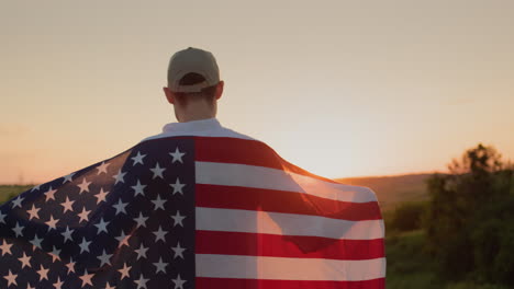 Young-male-farmer-with-usa-flag-on-his-shoulders-looks-at-sunrise-over-wheat-field