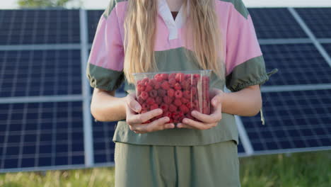 Teenage-girl-holds-a-plastic-container-with-fresh-raspberries.-The-panels-of-the-solar-power-plant-can-be-seen-in-the-background