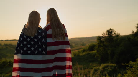 A-mother-with-a-teenage-daughter-with-an-American-flag-on-their-shoulders-look-at-the-sunset-over-a-picturesque-valley