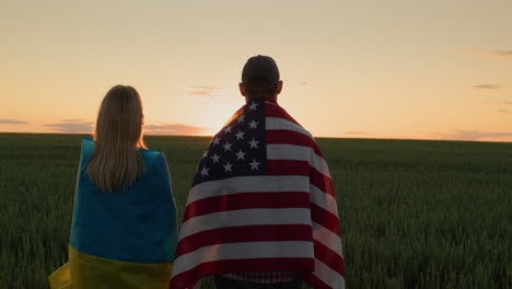 Ein-Mann-Und-Eine-Frau-Mit-Den-Flaggen-Der-Ukraine-Und-Der-USA-Stehen-Seite-An-Seite-Und-Betrachten-Den-Sonnenaufgang-über-Einem-Weizenfeld