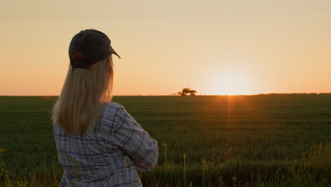 A-successful-woman-farmer-in-the-city-looks-at-her-wheat-field,-where-a-tractor-is-working-in-the-distance.-Silhouette-at-sunset