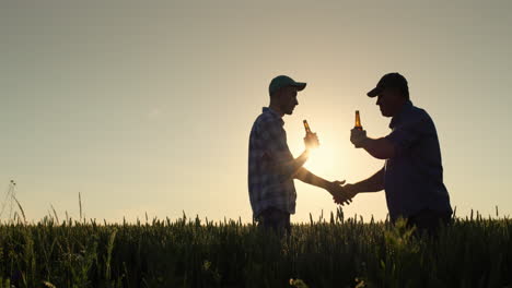 Silhouettes-of-two-men-clink-bottles-of-beer-and-shake-hands.-Standing-in-a-field-of-wheat-at-sunset
