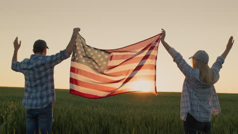 A-happy-couple-raises-up-the-US-flag,-standing-against-the-backdrop-of-a-wheat-field-at-sunset.