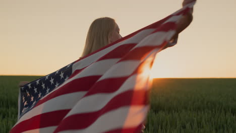 Handheld-shot:-A-woman-with-an-American-flag-on-her-shoulders-watches-the-sun-go-down-over-a-field-of-wheat.