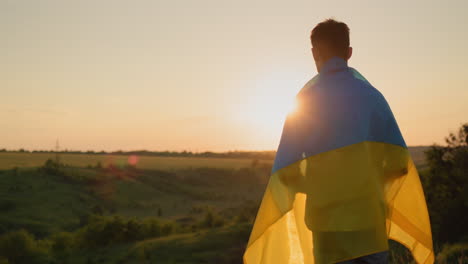A-young-man-with-the-flag-of-Ukraine-on-his-shoulders-looks-at-the-sunset-over-a-picturesque-valley