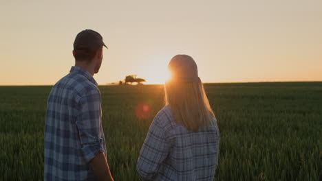 A-young-couple-of-farmers-watch-how-a-tractor-works-in-the-field.-Stand-side-by-side-against-the-backdrop-of-a-field-of-wheat-where-the-sun-is-setting