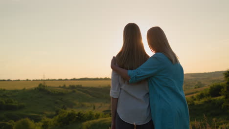 Mom-hugs-her-teenage-daughter,-watching-the-sunset-over-a-picturesque-valley.-Back-view