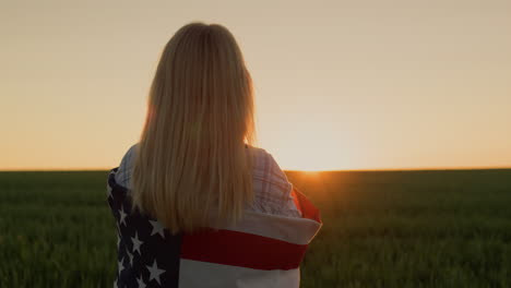 Eine-Frau-Mit-Einer-Amerikanischen-Flagge-Auf-Ihren-Schultern-Sieht-Zu,-Wie-Die-Sonne-über-Einem-Weizenfeld-Untergeht.