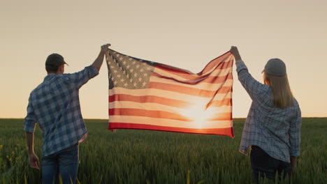 Dos-Agricultores-Felices-Izan-La-Bandera-Estadounidense-Sobre-Un-Campo-De-Trigo-Mientras-Se-Pone-El-Sol.-4-De-Julio---Concepto-Del-Día-De-La-Independencia-De-EE.UU.
