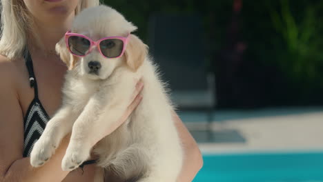 A-woman-in-a-bikini-holds-a-blonde-puppy-in-sunglasses-in-her-arms.-Summer-and-vacation-with-a-pet-concept