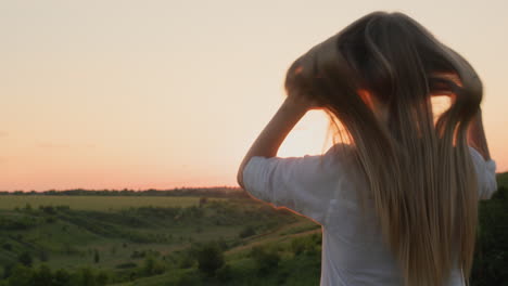 A-teenage-girl-touches-her-long-blond-hair,-stands-against-the-backdrop-of-a-picturesque-landscape-at-sunset.-Back-view