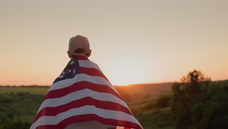 A-man-with-an-American-flag-on-his-shoulders-against-the-backdrop-of-a-picturesque-valley-where-the-sun-sets