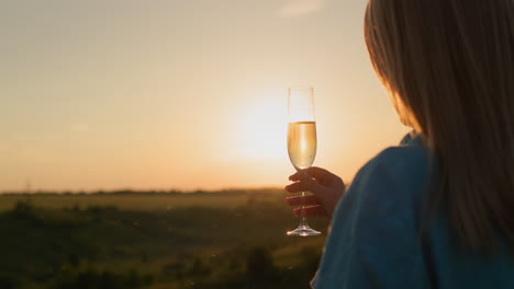 Back-view:-Successful-woman-with-a-glass-of-champagne-on-the-background-of-a-picturesque-valley-at-sunset