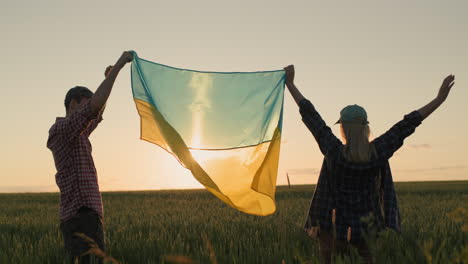Energisches-Ukrainisches-Paar,-Das-Bei-Sonnenuntergang-Die-Flagge-Der-Ukraine-über-Einem-Weizenfeld-Hisst