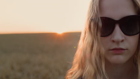 Portrait-of-a-teenage-girl-in-sunglasses-on-the-background-of-a-field-of-wheat-where-the-sun-sets