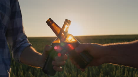 The-hands-of-two-men-with-bottles-of-beer-clink-against-the-background-of-a-field-of-wheat-where-the-sun-sets