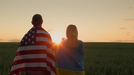 Couple-with-the-flags-of-Ukraine-and-the-USA-stand-side-by-side-and-look-at-the-sunrise-over-a-field-of-wheat