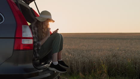 A-teen-girl-sits-in-the-trunk-of-a-car,-uses-a-smartphone.-Against-the-backdrop-of-a-rural-landscape-where-the-sun-sets.-4k-video