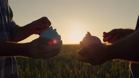 A-couple-of-farmers-put-money-in-a-piggy-bank,-stand-against-the-backdrop-of-a-wheat-field-at-sunset.-Investments-and-savings-in-agriculture