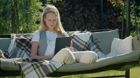 A-teenage-girl-uses-a-tablet,-sits-on-a-garden-swing-in-the-backyard-of-a-house.