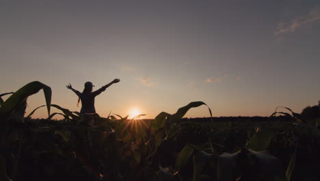 A-young-female-farmer-raises-her-hands-over-a-field-of-corn-where-the-sun-is-setting