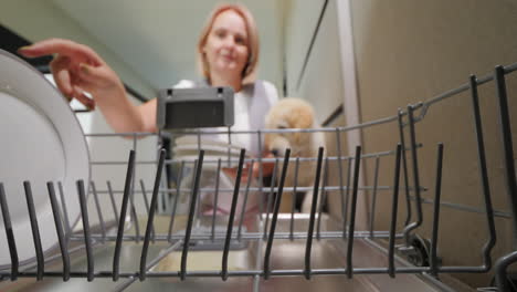 Cute-woman-takes-clean-plates-from-the-dishwasher.-Her-puppy-is-next-to-her