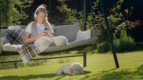 A-teenage-girl-is-resting-on-a-garden-swing,-using-a-smartphone.-Her-puppy-sits-next-to-her