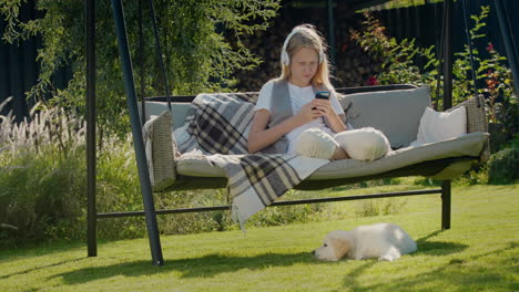 Una-Adolescente-Descansa-En-Un-Columpio-De-Jardín,-Usando-Un-Teléfono-Inteligente.-Su-Cachorro-Se-Sienta-A-Su-Lado