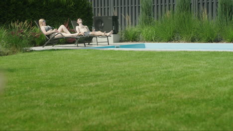 A-young-couple-is-relaxing-by-the-pool-in-the-backyard-of-the-house.-In-the-foreground,-a-well-groomed-green-lawn