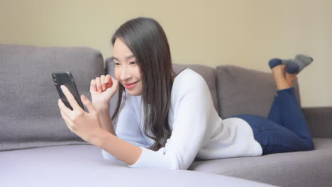Young-Happy-Asian-Woman-Lying-on-Couch-and-Looking-at-Smartphone,-Social-Network-or-Selfie-Picture-Concept,-Full-Frame-Slow-Motion
