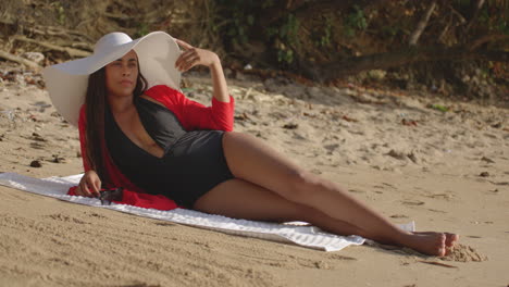 Latin-Model-Woman-on-Beach-Wearing-Hat-Lying-Down-on-her-side-puts-on-Sunglasses-facing-camera