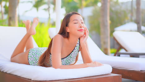 Young-Asian-girl-in-spotty-monokini-lying-on-her-belly-on-a-comfortable-deck-chair-in-a-gorgeous-tropical-resort-smiling,-daytime-slow-motion