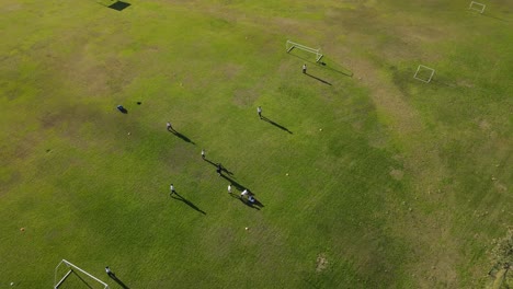 Aerial-drone-shot-showing-team-of-children-playing-football-soccer-on-green-lawn-during-summer