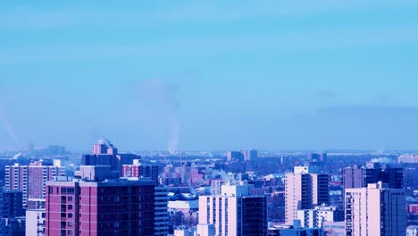 4k-Time-Lapse-low-rise-apartment-buildings-with-steam-rising-from-heaters-on-a-windy-sunny-winter-afternoon-in-the-foreground-and-factory-structures-in-the-background-pumping-out-white-thick-smoke