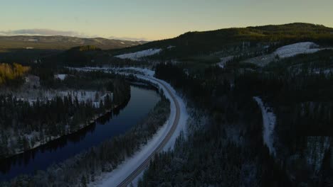 Curved-Country-Road-Along-Calm-River-and-Vast-Forest,-Winter-Season,-Aerial