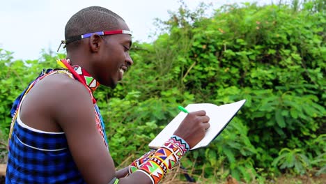 African-male,-smiling-while-writing-notes-in-a-hes-diary,-wearing-tribal-gear