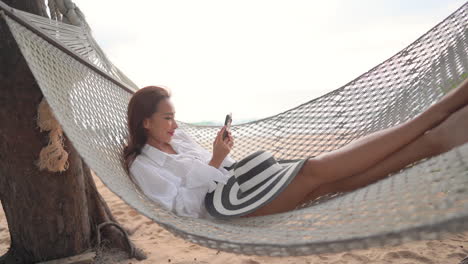 Young-asian-woman-laying-on-beach-hamcock-and-looking-at-smartphone-with-smile