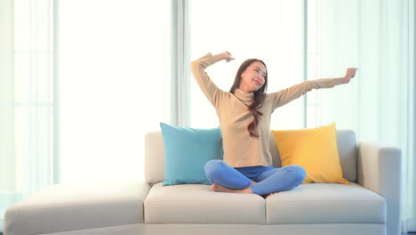 Asian-young-woman-stretching-her-arms-over-the-head-while-sitting-on-a-sofa-having-her-legs-crossed-wearing-casual-clothes-daytime--static-shot
