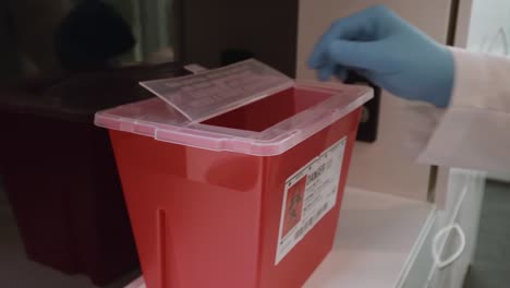 Gloved-hand-throwing-empty-vial-and-contaminated-needle-in-sharps-disposal-container