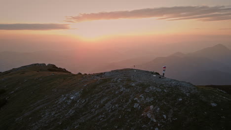 Drohnenaufnahme-Vom-Gipfel-Des-Berges-Bei-Sonnenaufgang,-Wanderer-Mit-Einer-Slowenischen-Flagge-Auf-Dem-Pol,-Die-In-Der-Brise-Flattert