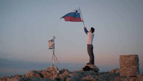Mann-Steht-Auf-Einem-Felsen-Und-Hält-Einen-Wanderstock-In-Der-Luft-Mit-Befestigter-Slowenischer-Flagge