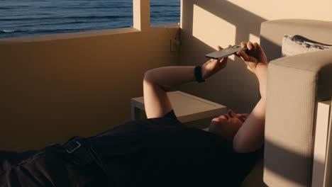Boy-Lying-Down-Holding-Phone-in-hands,-Hotel-Balcony-in-Sunlight-with-Sea-View,-Hawaii