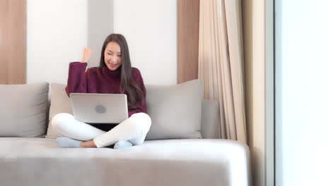 Happy-Asian-Female-Working-on-Laptop-in-a-Living-Room-With-Winning-Hand-Gesture