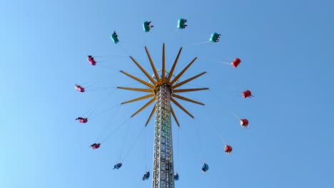 Vertical-colorful-chain-fair-swing-spinning-people-around,-symmetrical-minimal-background-blue-sky,-static