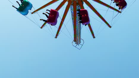Low-angle-view-of-colorful-chain-fair-swing-spinning-people-around,-minimal-background-blue-sky,-static
