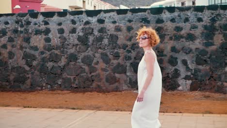 A-redhead-woman-dressed-in-white-walking-along-a-black-wall-in-a-windy-day