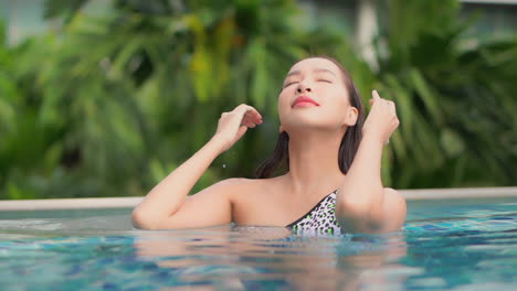 Beauty-shot,-Asian-woman-touching-hair-and-smoothing-out-hair-in-swimming-pool-on-a-sunny-day,-Face-close-up-slow-motion-front-view